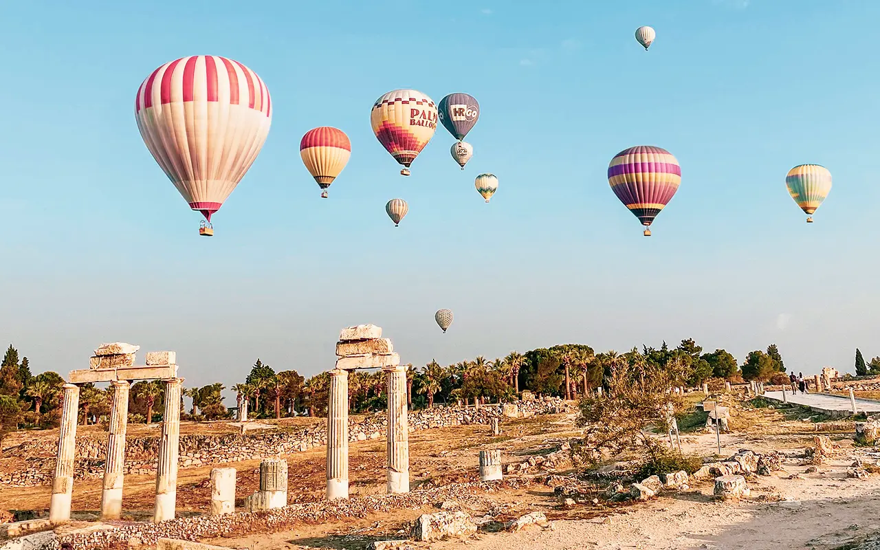 Balloons drifting over the ancient Hierapolis columns next to Pamukkale's travertines during a sunrise flight.