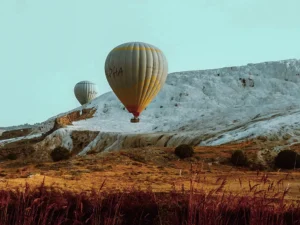 Balloon skimming the edge of Pamukkale's travertine cliffs at low altitude, autumn reeds in the foreground.