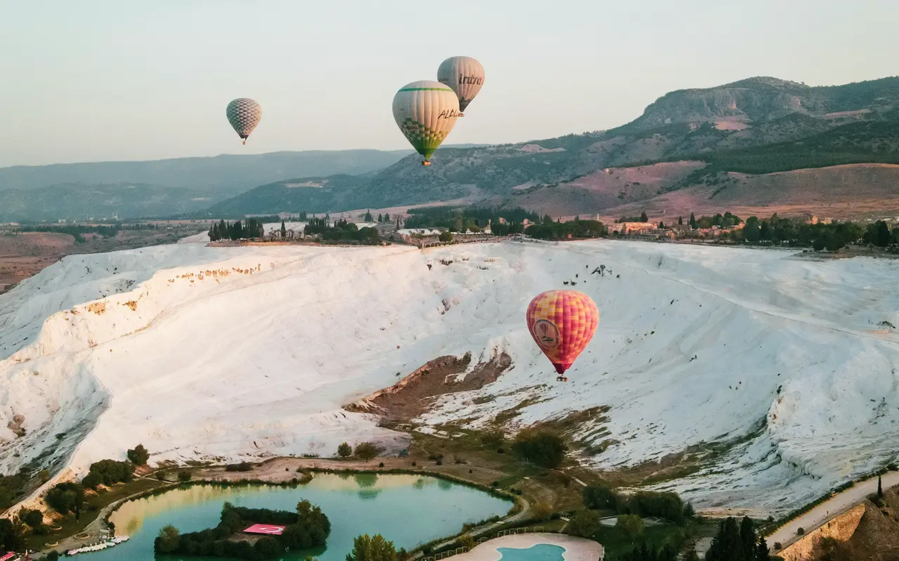Balloons floating over Pamukkale's white travertine terraces at low altitude, a natural pool visible in the foreground.
